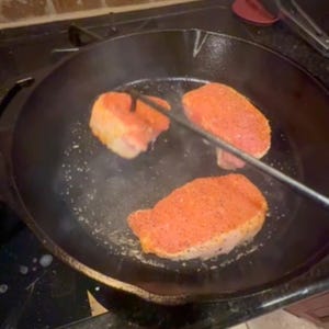Puede incluir: Tres chuletas de cerdo sazonadas cocin&aacute;ndose en una sart&eacute;n de hierro fundido negro sobre una estufa. Una chuleta se sostiene con pinzas de metal. Las chuletas est&aacute;n cubiertas con una mezcla de especias rojas y parecen chisporrotear.