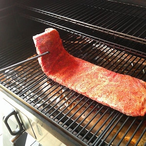 Puede incluir: Una gran costilla sazonada en una parrilla. Las costillas est&aacute;n cubiertas con una vibrante mezcla de especias rojas y se est&aacute;n cocinando en una rejilla de metal. La parrilla est&aacute; abierta y las costillas se cocinan a fuego indirecto.