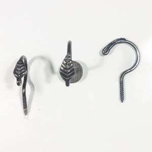 May include: Three black metal hooks. Two have a leaf design, one with a screw base and the other with a flat round base. The third hook is a simple curved design with a screw base. All are against a white background.