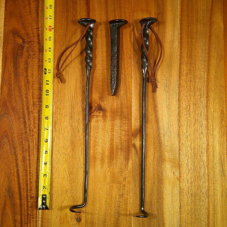 Puede incluir: Tres herramientas de metal negro para fogatas y un clavo grande se muestran sobre una superficie de madera. Dos herramientas tienen mangos retorcidos y lazos de cuero. Las herramientas miden aproximadamente 43 cm de largo.