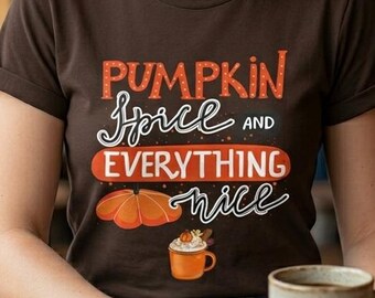 May include: A brown t-shirt with the text "Pumpkin Spice and Everything Nice" in orange and white. The shirt features an illustration of a pumpkin and a mug of coffee. The person is holding a mug.