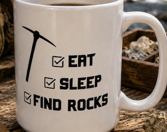 May include: A white ceramic mug featuring a black graphic of a pickaxe and a checklist with the text EAT, SLEEP, FIND ROCKS. The mug has a standard handle and is positioned among various rocks, a brown journal, a magnifying loupe, and a backpack.