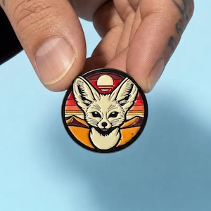 May include: A circular enamel pin featuring a cream-coloured fennec fox head centred against a desert landscape background. The design includes a setting sun, dark red and orange horizontal sky stripes, and orange sand dunes with a black border.