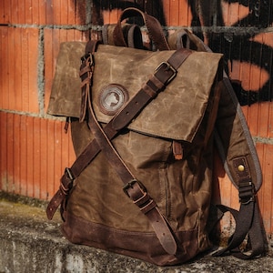 May include: A brown canvas backpack with leather straps and buckles. The backpack has a flap closure and a large pocket on the front. The backpack has a logo on the front that says "Woofwalker".