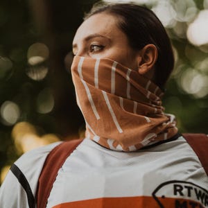 May include: A tan and white striped neck gaiter worn by a person. The gaiter covers the lower face and neck. The person is wearing a white and orange athletic shirt with a logo on the sleeve. The background is blurred green.