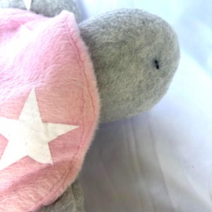 May include: A plush gray turtle with a pink shell. The shell features white star accents. The turtle's head is gray, and the plush toy is set against a white background. The toy is soft and cuddly.