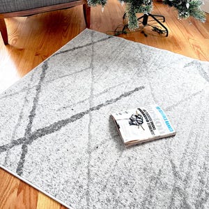 以下が含まれることがあります： 濃い灰色の線で幾何学模様が描かれた、明るい灰色のラグ。 「Boston」というタイトルの雑誌がラグの上にあります。 ラグは木製の床にあり、灰色のアームチェアとクリスマスツリーの近くにあります。