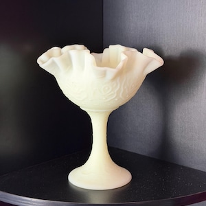 Puede incluir: Un cuenco de cristal translúcido de color amarillo pálido con un borde ancho y ondulado y una base de pedestal. La superficie exterior del cuenco y el tallo están decorados con un patrón floral en relieve que cubre todo el cuerpo de cristal.