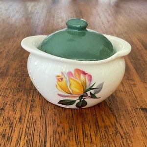 May include: A small white ceramic sugar bowl with a green lid and a yellow rose design.