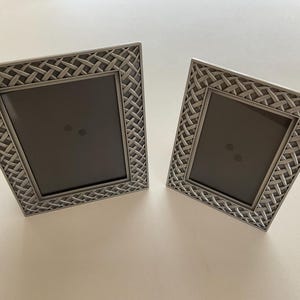 May include: Two silver-coloured picture frames with a woven pattern border. The frames have a rectangular opening for photos and are angled slightly towards the viewer. The frames are made of metal and are suitable for displaying photographs.