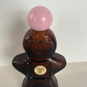 May include: A brown glass bottle shaped like a gingerbread person with a pink ball stopper. The bottle has a label that reads "Delicate Daisies Cologne 1/2 fl oz."