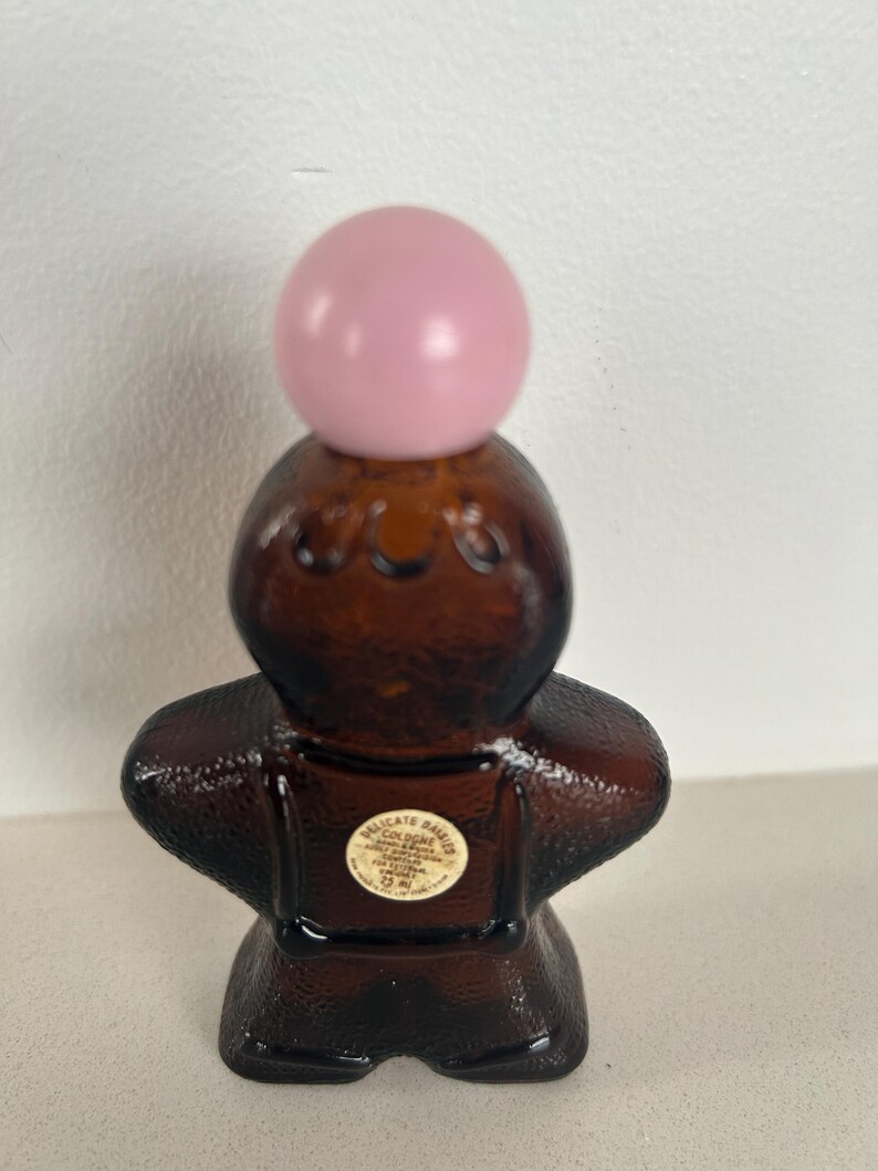 May include: A brown glass bottle shaped like a gingerbread person with a pink ball stopper. The bottle has a label that reads "Delicate Daisies Cologne 1/2 fl oz."