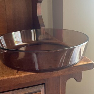 May include: A clear, brown glass baking dish with a rounded bottom and slightly flared sides. The dish is sitting on a wooden surface.