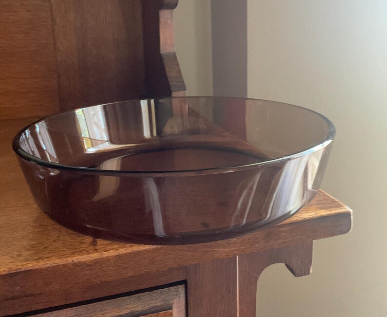May include: A clear, brown glass baking dish with a rounded bottom and slightly flared sides. The dish is sitting on a wooden surface.