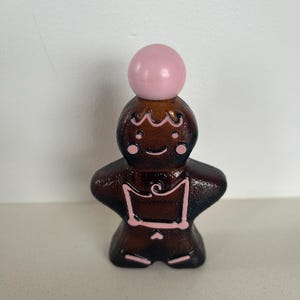 May include: A brown glass bottle shaped like a gingerbread person with pink details. The bottle has a pink ball shaped stopper on top.