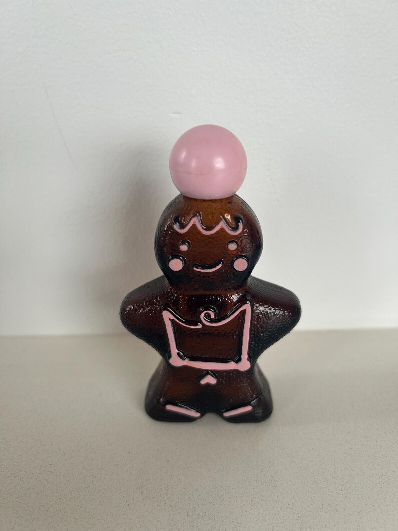 May include: A brown glass bottle shaped like a gingerbread person with pink details. The bottle has a pink ball shaped stopper on top.