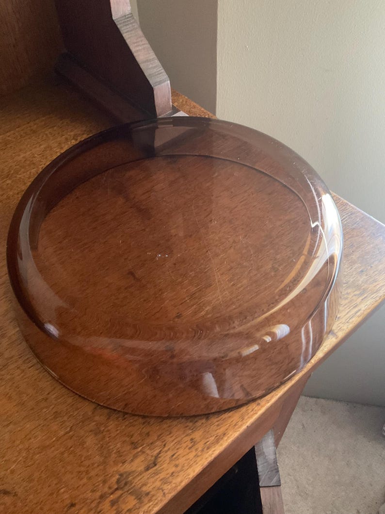 May include: A clear glass dome with a brown tint. The dome is sitting on a wooden surface.