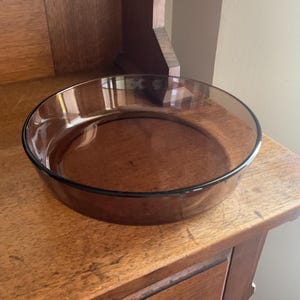 May include: A round, clear, brown glass baking dish. The dish is empty and sitting on a wooden surface.