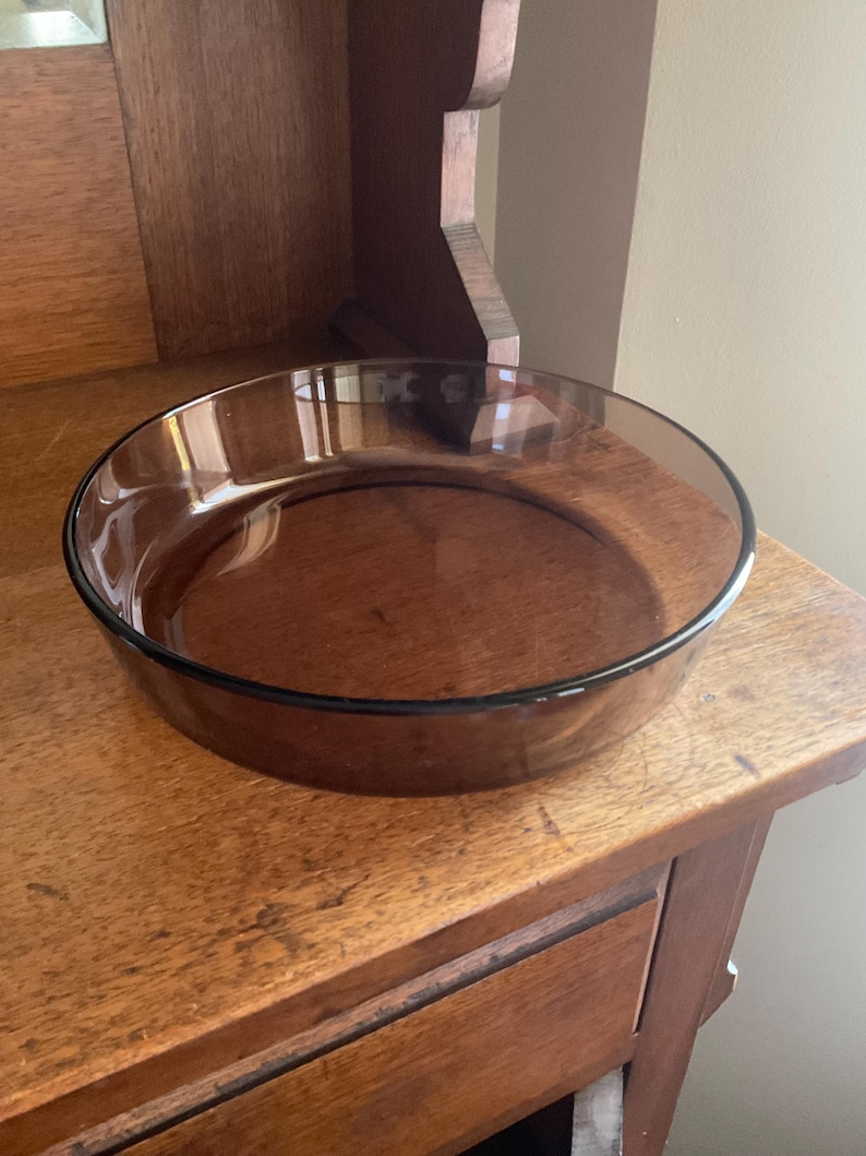 May include: A round, clear, brown glass baking dish. The dish is empty and sitting on a wooden surface.