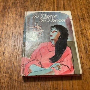 May include: A vintage book cover titled "To Dance, To Dream." The cover shows an illustration of a woman in a pink dress and a ballerina. The book has a worn appearance, with visible signs of age and use.