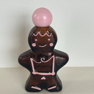 May include: A brown glass gingerbread man bottle with a pink plastic stopper. The bottle has pink icing details.