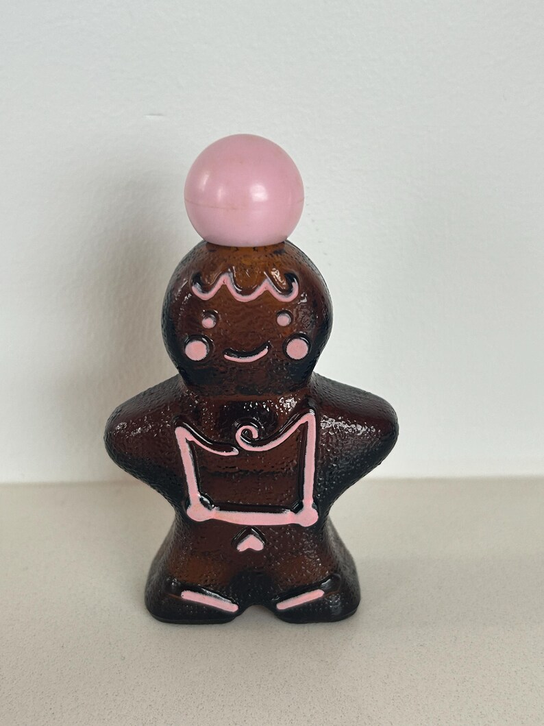 May include: A brown glass gingerbread man bottle with a pink plastic stopper. The bottle has pink icing details.