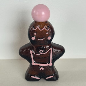 May include: A brown glass bottle shaped like a gingerbread person with a pink plastic ball on top. The bottle has white icing details and a pink heart on the front.