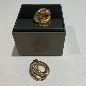 May include: A gold-coloured brooch with an oval design, featuring a textured surface and small decorative accents. The brooch is displayed on a dark surface, with a matching brooch placed below. The jewelry piece has a polished finish.