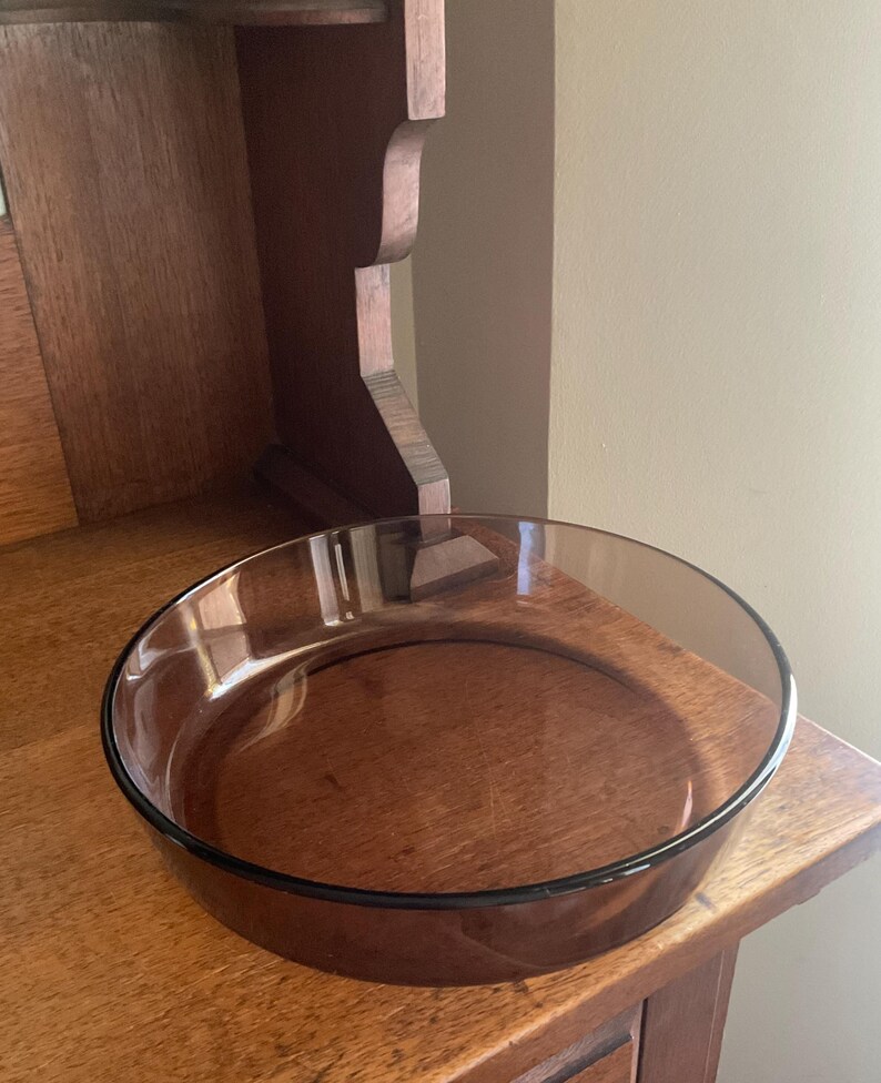 May include: A round, clear glass baking dish with a brown tint. The dish is sitting on a wooden surface.