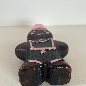 May include: A brown glass bottle shaped like a person with pink painted details. The bottle has a round, flat bottom.