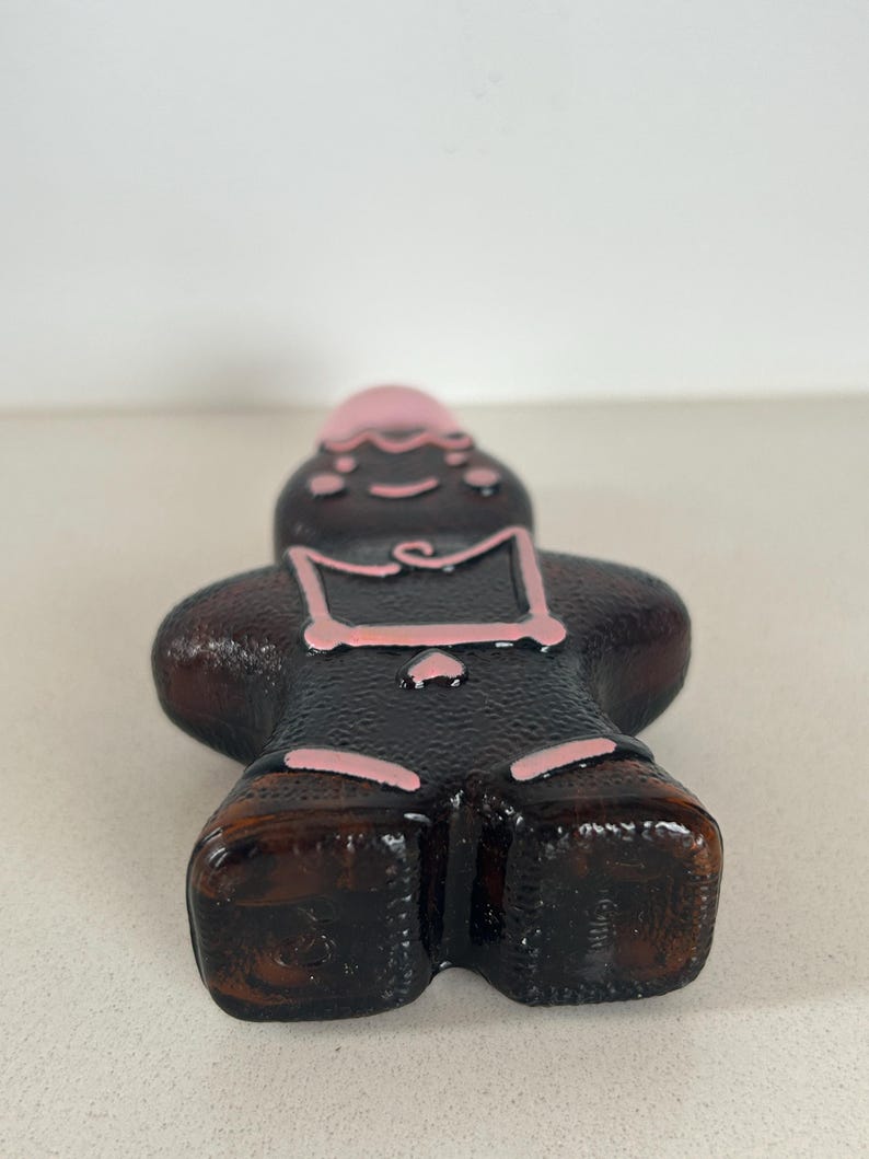 May include: A brown glass bottle shaped like a person with pink painted details. The bottle has a round, flat bottom.