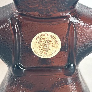 May include: A brown glass bottle shaped like a woman wearing a dress and a bonnet. The bottle has a white label that reads "Delicate Daisies Cologne" and "Handle Under Adult Supervision". The label also states "Contents for External Use Only" and "25 ml".