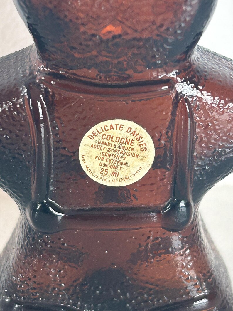 May include: A brown glass bottle shaped like a woman wearing a dress and a bonnet. The bottle has a white label that reads "Delicate Daisies Cologne" and "Handle Under Adult Supervision". The label also states "Contents for External Use Only" and "25 ml".