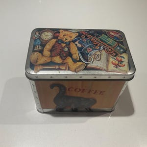 May include: A rectangular metal coffee tin with a decorative lid. The lid features a teddy bear, clock, train, and other vintage-style illustrations. The front of the tin has a black cat silhouette and the word "COFFEE".