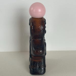 May include: A brown glass bottle with a pink plastic stopper shaped like a ball. The bottle has a textured surface and is shaped like a person.