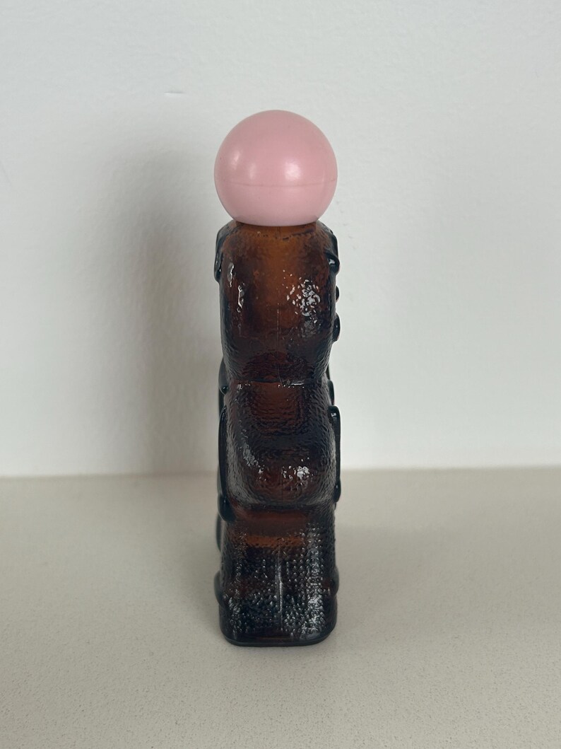 May include: A brown glass bottle with a pink plastic stopper shaped like a ball. The bottle has a textured surface and is shaped like a person.