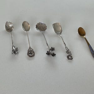May include: Five decorative spoons with ornate designs. The spoons feature various styles, including floral and leaf motifs, and a small spoon with a blue and gold handle. The spoons are made of silver and gold.