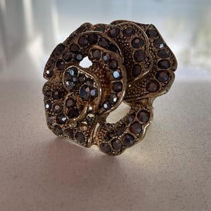 May include: A gold-coloured ring in the shape of a rose, adorned with dark, sparkling gemstones. The ring's design features detailed petals and a textured band, creating a vintage or antique aesthetic. The gemstones are closely set, enhancing the ring's brilliance.