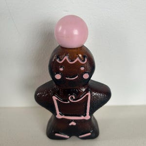 May include: A brown glass bottle shaped like a gingerbread person with pink icing details and a pink ball on top.
