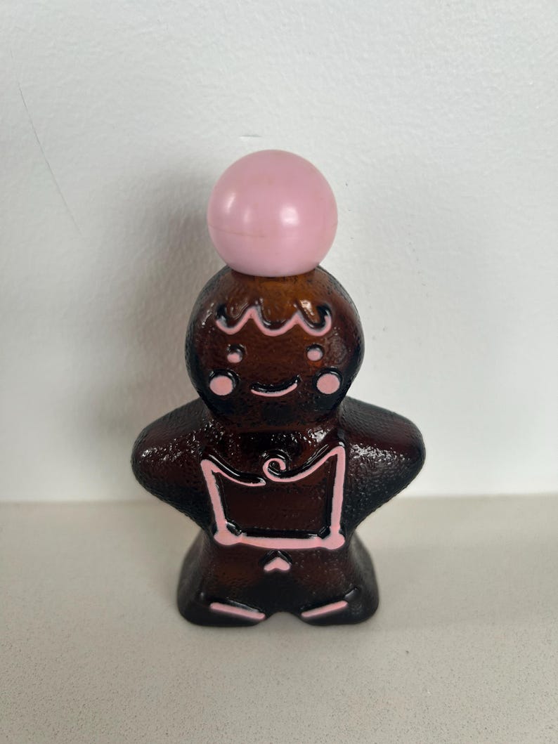 May include: A brown glass bottle shaped like a gingerbread person with pink icing details and a pink ball on top.