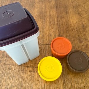 May include: A collection of vintage Tupperware containers. The set includes a clear, rectangular container with a dark purple lid, and three round lids in yellow, orange, and brown. The containers are arranged on a wooden surface.