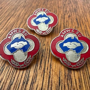 May include: Three vintage AMW & SU union pins. Each pin features a red and silver quatrefoil design with a blue center depicting a handshake over a map. The pins are on a wooden surface.