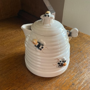 Peut inclure: Une crémier ou un sucrier en céramique blanche en forme de ruche avec des détails d'abeilles. Le couvercle est en forme d'abeille avec des yeux noirs.