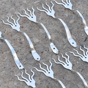 Könnte beinhalten: Eine Sammlung von 16 silberfarbenen Metallgabeln, die zu einem einzigartigen Design gebogen und geformt wurden. Die Gabeln sind in Reihen auf einer grauen Betonfläche angeordnet.