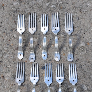 Peut inclure: Un ensemble de 12 fourchettes en argent disposées en deux rangées de six, les dents vers le haut. Les fourchettes sont disposées de manière symétrique sur une surface en béton gris.