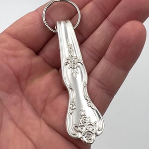 Puede incluir: Un llavero plateado con un mango de cuchara con detalles florales. El mango de la cuchara está unido a un llavero plateado. El diseño incluye flores y hojas en relieve. Un accesorio único y artesanal.