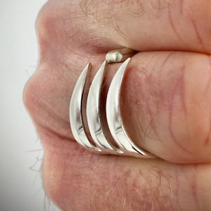 May include: A silver ring with three curved bands, worn on a person's finger.