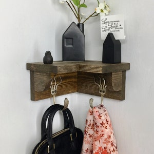Peut inclure: Une étagère d'angle en bois avec deux crochets, un vase en céramique noire en forme de maison avec des fleurs blanches, une figurine de maison en céramique noire et une carte blanche avec le texte "Live every moment, Laugh every day, Love beyond words". Un sac à main noir et une écharpe florale rose et rouge sont accrochés aux crochets.