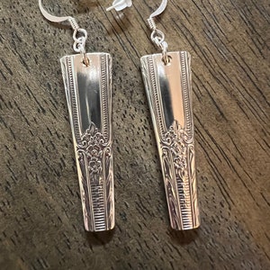 May include: A pair of silver earrings made from vintage silverware. The earrings are long and narrow, with a decorative design on the front. The earrings are attached to silver hooks.