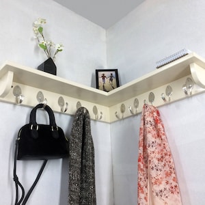 Puede incluir: Estantería esquinera color crema con múltiples ganchos plateados para colgar objetos. Un bolso negro, una bufanda estampada y una bufanda floral cuelgan de los ganchos. La estantería también contiene una pequeña foto enmarcada, un bloc de notas y un jarrón negro con flores blancas.